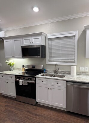 A modern kitchen with white cabinetry, a stainless steel microwave, a built-in oven, a dishwasher, and a window with blinds.