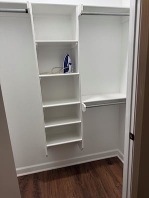 Een inloopkast met witte planken en een kledingstang, met een blauw en wit ijzeren strijkobject op een plank.