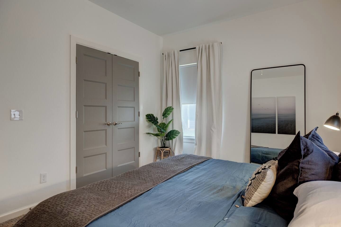 Une chambre à coucher avec un lit, une lampe de chevet, un miroir, une fenêtre avec des rideaux et une porte.