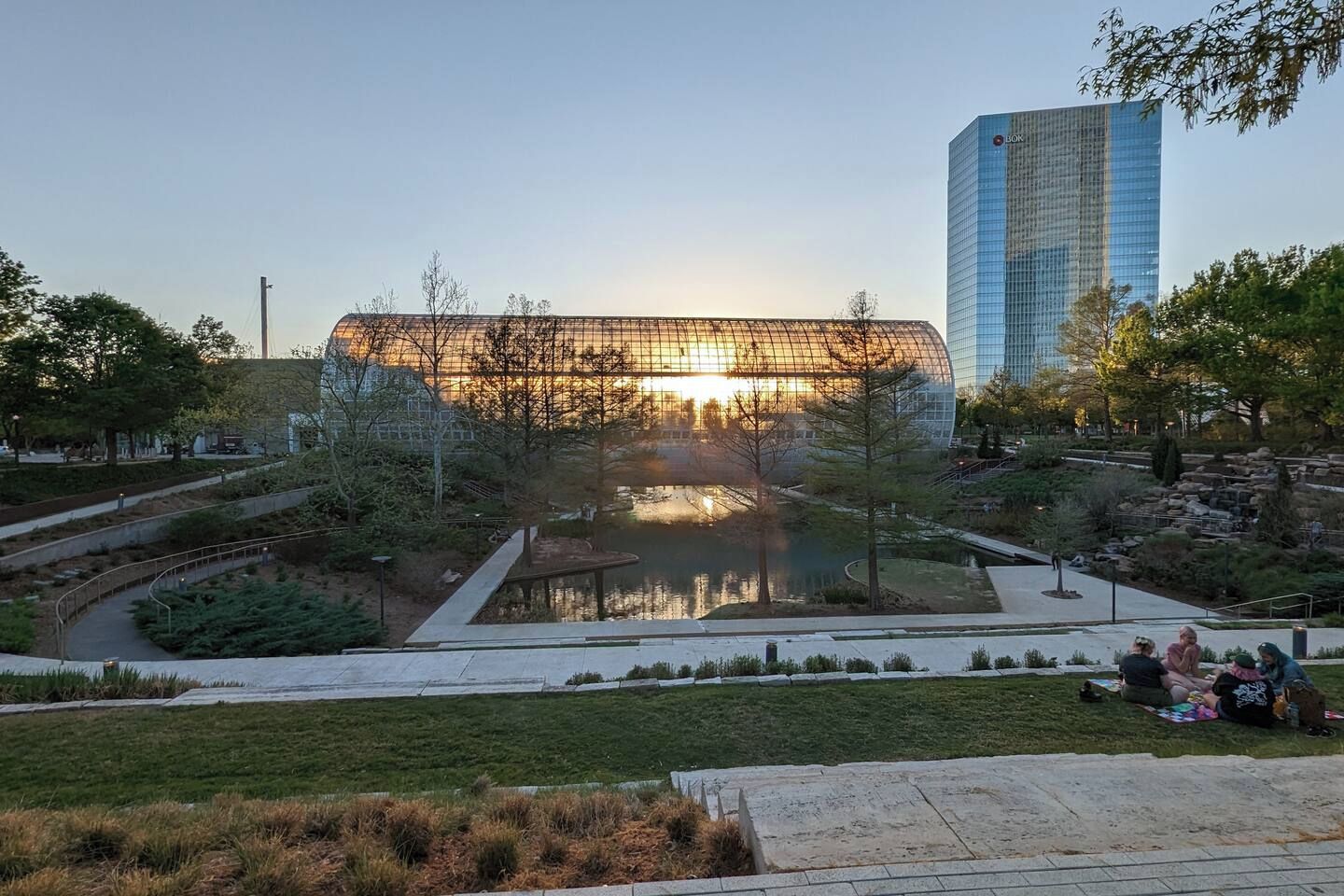 A modern building with a glass facade, a reflective water feature in front, and a park area with pathways and greenery.