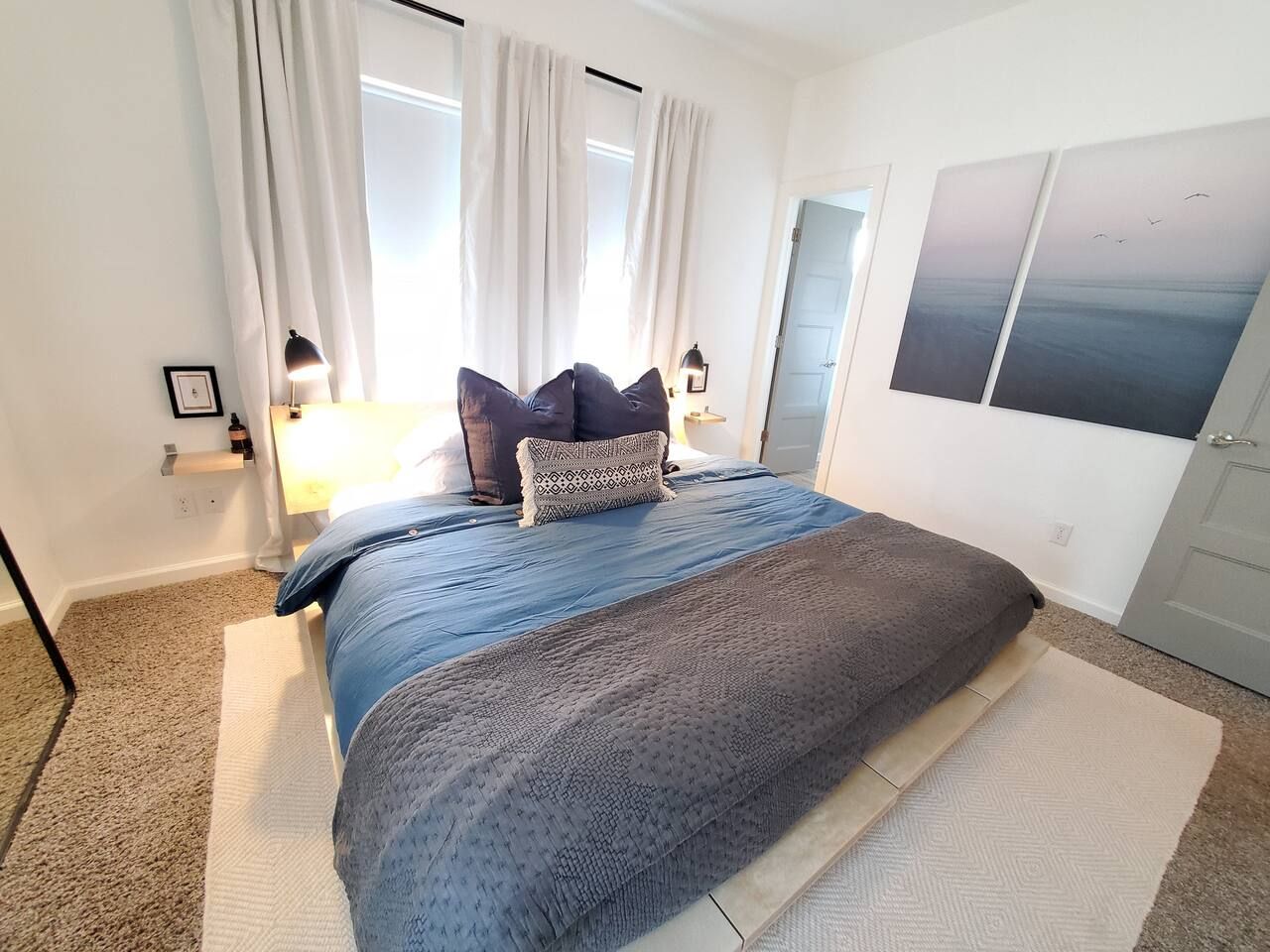 Un dormitorio con cama, cortinas, una mesita de noche, lámpara y cuadros en la pared.