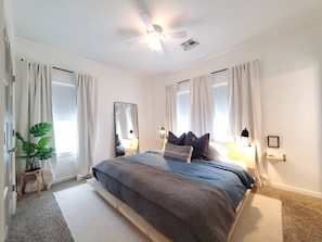 Een slaapkamer met een bed, gordijnen, een plafondventilator, een spiegel en een bedlampje.