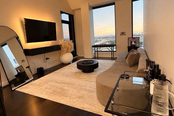 A modern living room with a large flat-screen TV, a round coffee table, a sofa, and a view of a cityscape through large windows.