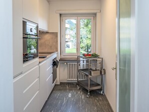 Eine kompakte Küche mit weißen Schränken, einem eingebauten Backofen, einem Fenster mit Blick auf Grünflächen und einem kleinen Metallregal mit Küchenutensilien.