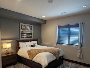 Un dormitorio con cama, mesita de noche, lámpara, ventana con persianas y un cuadro en la pared.