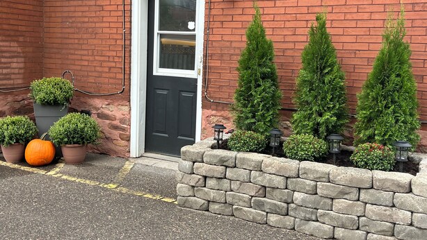 Una casa de ladrillo con puerta negra, plantas en macetas y un muro de piedra.