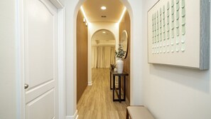 Un couloir doté d’un plancher en bois, d’une porte blanche, d’une table sur laquelle se trouvent un vase et un miroir, ainsi que d’un panneau décoratif fixé au mur.