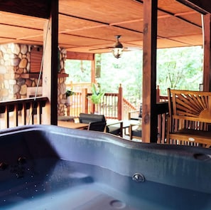 A hot tub under a wooden pergola with a stone wall backdrop, outdoor furniture, and a view of greenery. - Heated Pool/Cinema/Hot Tub/Game Room/Massage Chair (Broken Bow)