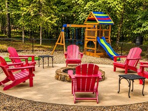 Ein Spielplatz mit Rutsche, Schaukeln und einem Spielhäuschen. Rote Adirondack-Stühle und eine Feuerstelle sind ebenfalls vorhanden.