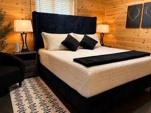 Ein Bett mit dunklem Kopfteil, weißen und schwarzen Kissen sowie einer schwarzen Tagesdecke.