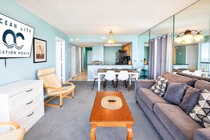 Ein modernes Wohnzimmer mit einer Couch, einem Couchtisch und einem Essbereich mit Stühlen. Im Hintergrund ist eine Küche zu sehen.