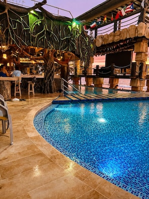An indoor swimming pool with a tiled edge, surrounded by a bar area with stools and a wooden structure overhead.