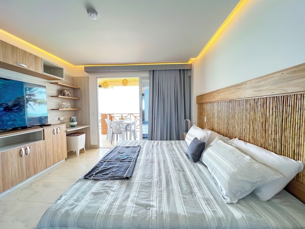 A modern hotel room with a large bed, a flat-screen TV, wooden cabinets, and a balcony with a table and chairs.