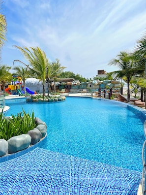 A clear blue swimming pool with a tiled edge, surrounded by palm trees and a play area with slides.