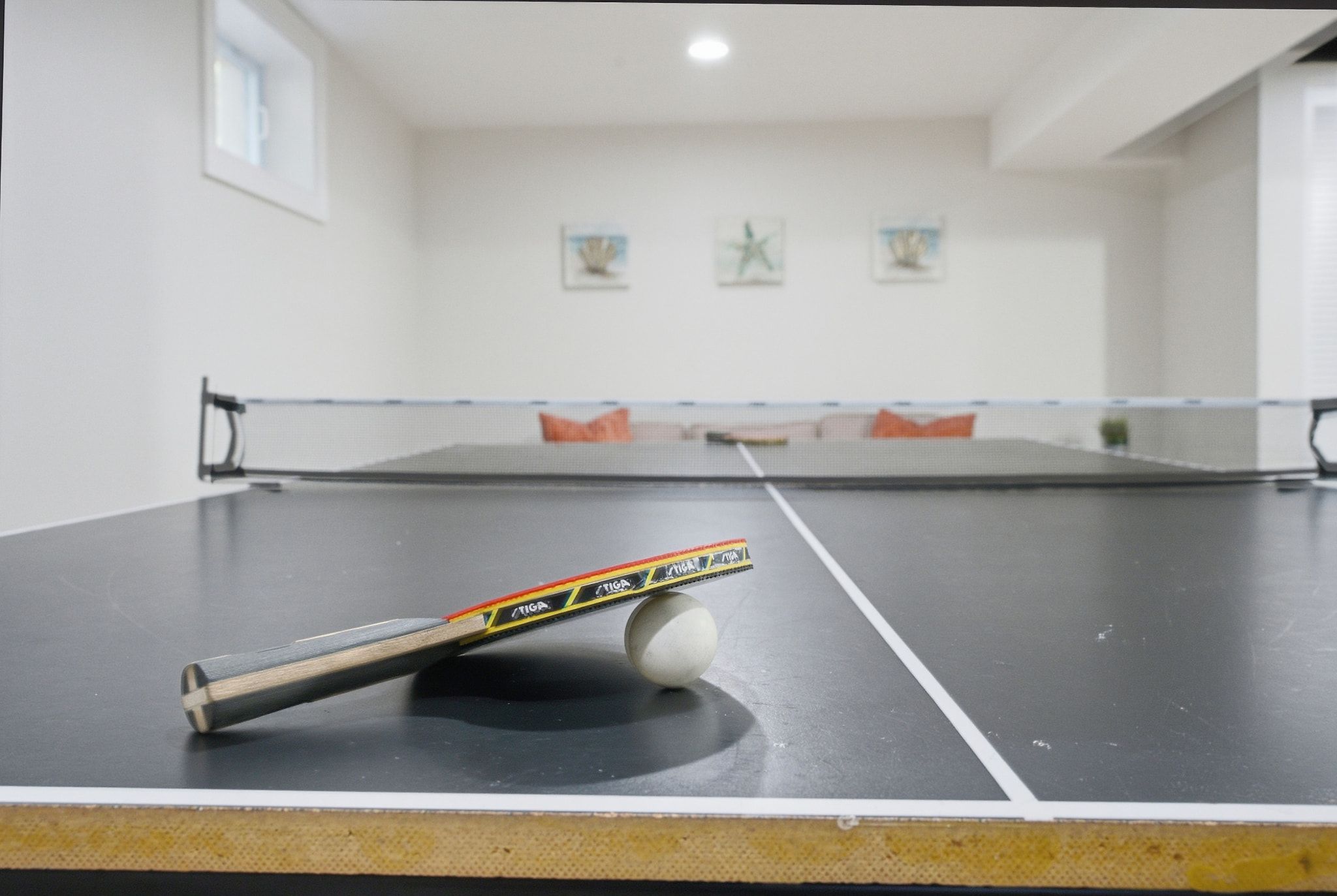 Une table de ping-pong avec une balle et une raquette au premier plan, un canapé avec des coussins à l’arrière-plan, et un mur orné de tableaux encadrés.