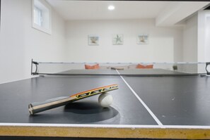 Una mesa de ping pong con una pelota y una raqueta en primer plano, un sofá con cojines al fondo y una pared con cuadros enmarcados.