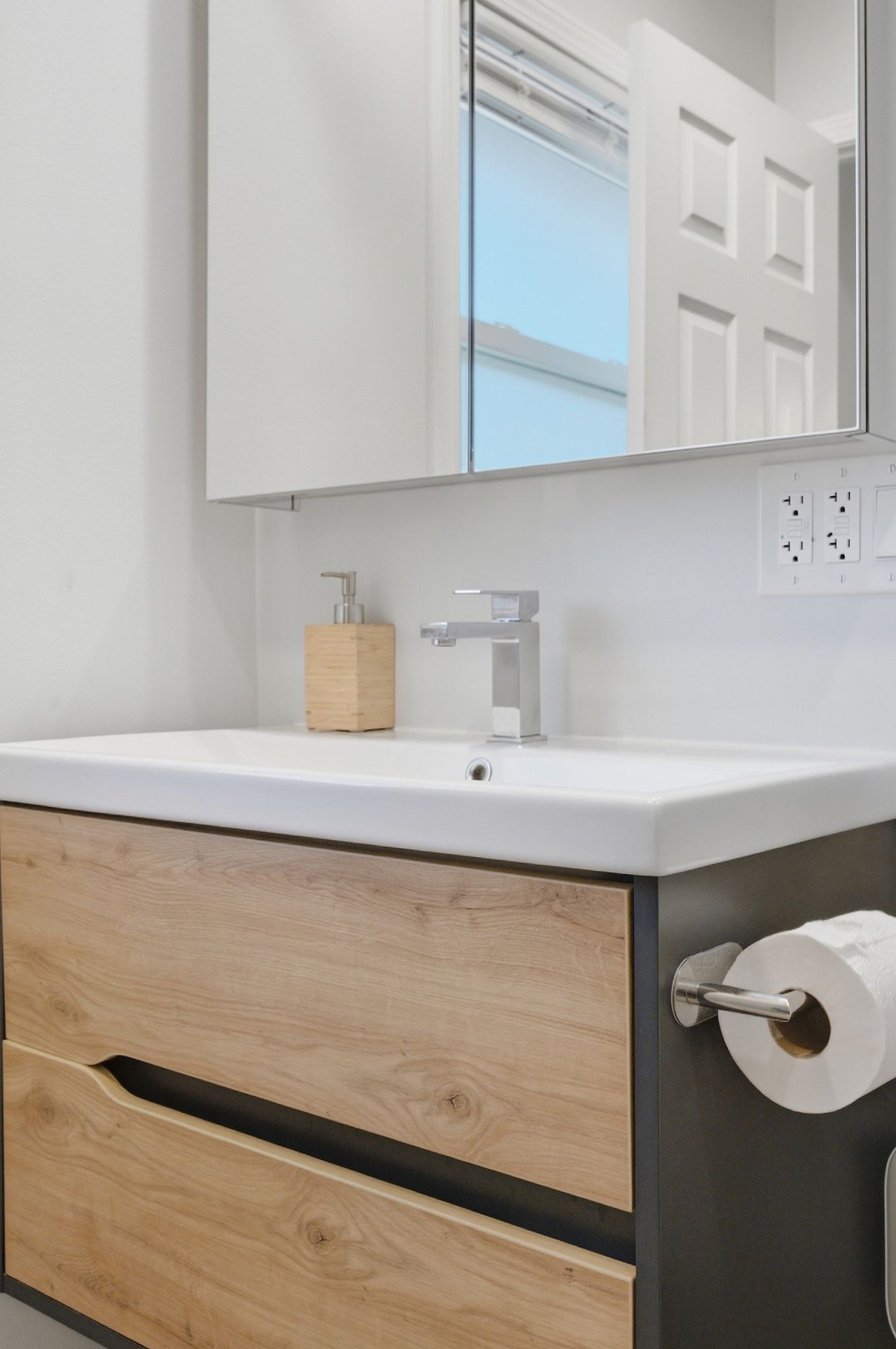 A bathroom with a white sink, wooden cabinet, and a mirror reflecting a door and window.
