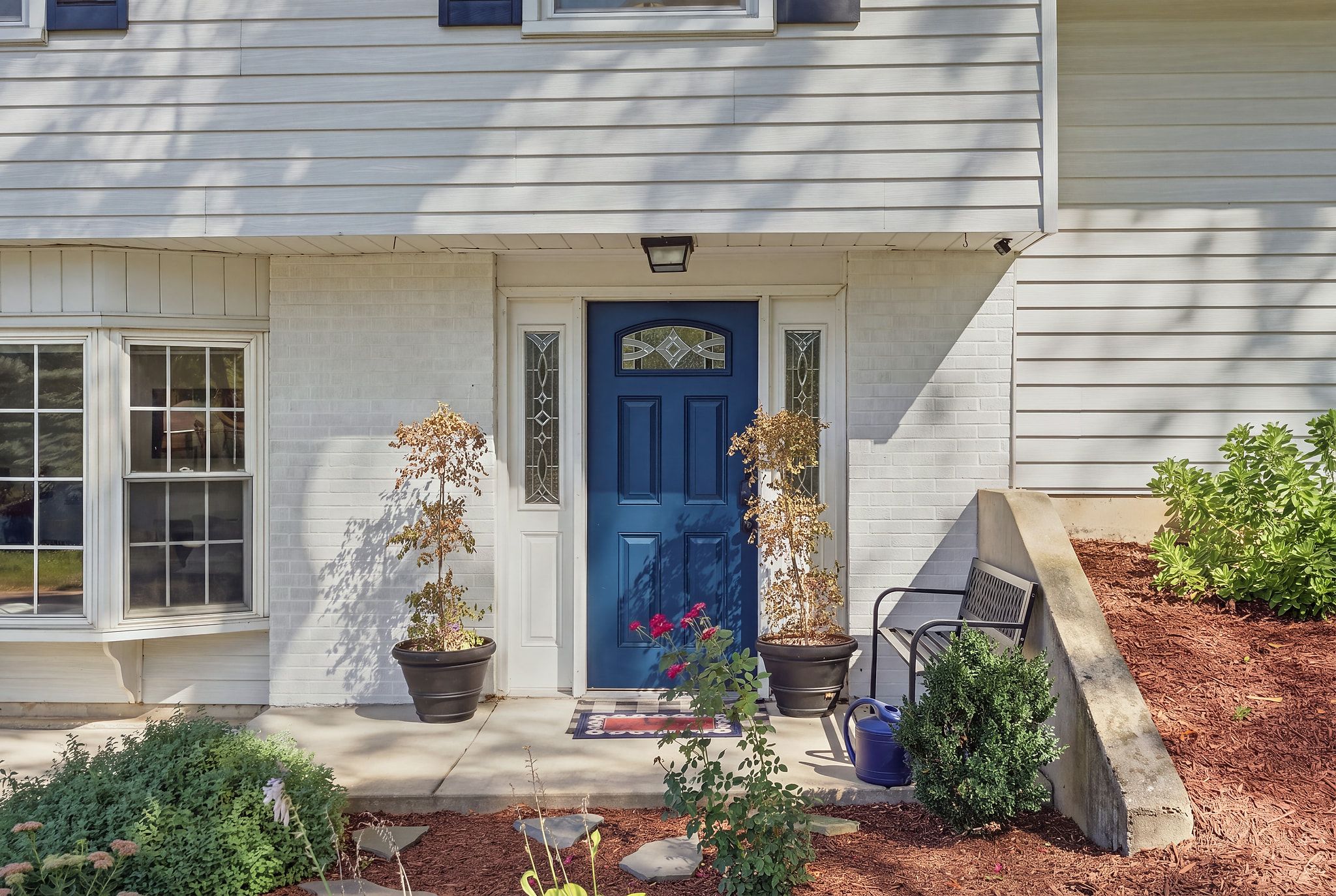 Une maison avec une porte d’entrée bleue, des plantes en pot, un banc et un petit jardin.