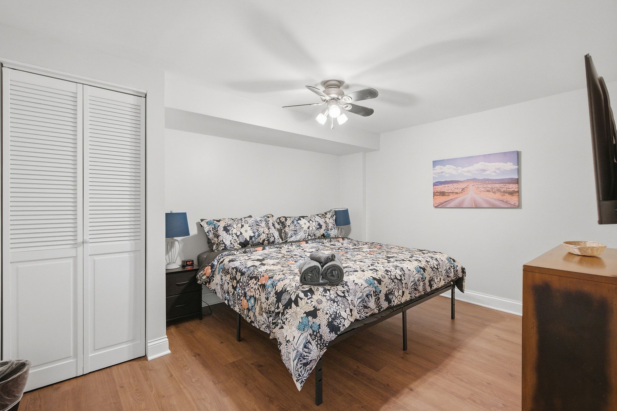 Une chambre à coucher comprenant un lit, un ventilateur de plafond, une table de chevet, une lampe, un téléviseur fixé au mur et un tableau.