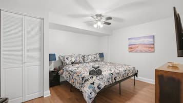 Une chambre à coucher comprenant un lit, un ventilateur de plafond, une table de chevet, une lampe, un téléviseur fixé au mur et un tableau.
