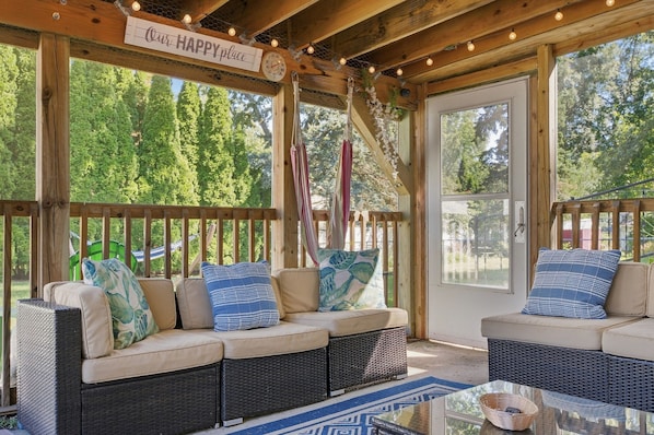 A covered outdoor seating area with wicker furniture, a sign reading 'Our Happy Place,' and a view of trees through large windows.