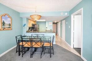 Uma sala de jantar com mesa e cadeiras de vidro, cozinha ao fundo e um corredor que leva a uma porta.