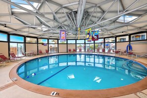 Uma grande piscina interior com água azul límpida, rodeada por espreguiçadeiras e camas de banho, sob um teto alto com vigas de metal.