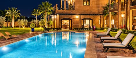 Una piscina con lettini, un edificio con finestre ad arco e palme.
