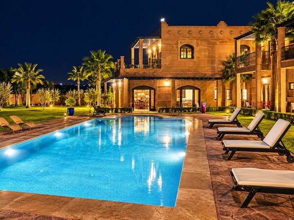 A swimming pool with lounge chairs, a building with arched windows, and palm trees.