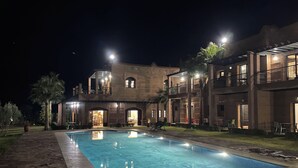 Een nachtzicht van een hotel met een zwembad, terras en een goed verlichte ingang.