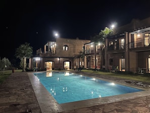 Vista noturna de um hotel com piscina, área de estar externa e entrada bem iluminada.