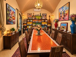 A dining area with a long table, chairs, a chandelier, bookshelves, and artwork.