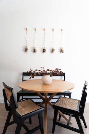 Una mesa redonda de madera con un jarrón blanco y una rama con hojas marrones. Cuatro sillas negras con asientos de mimbre la rodean.