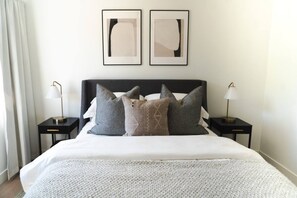 A neatly made bed with a white comforter and multiple pillows, flanked by two bedside tables with lamps, in a room with a plain wall and two framed abstract artworks above the bed.