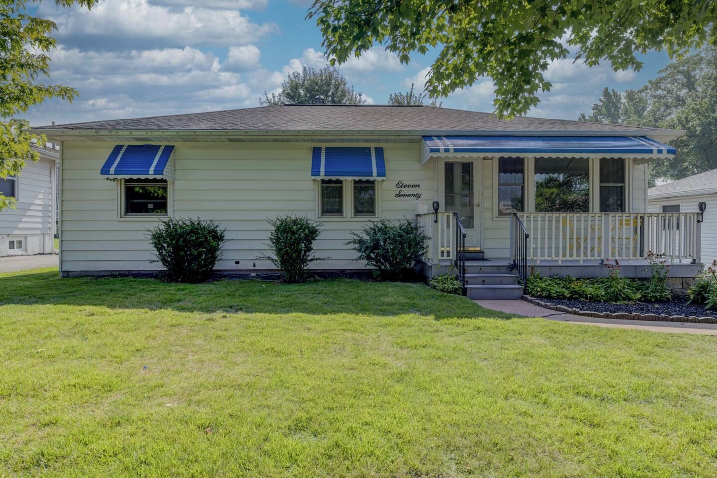 A single-story house with a front porch, two windows with blue awnings, and a small front yard.