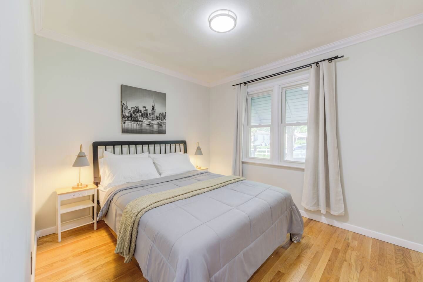 A bedroom with a bed, bedside lamps, a nightstand, a window with curtains, and a cityscape painting on the wall.