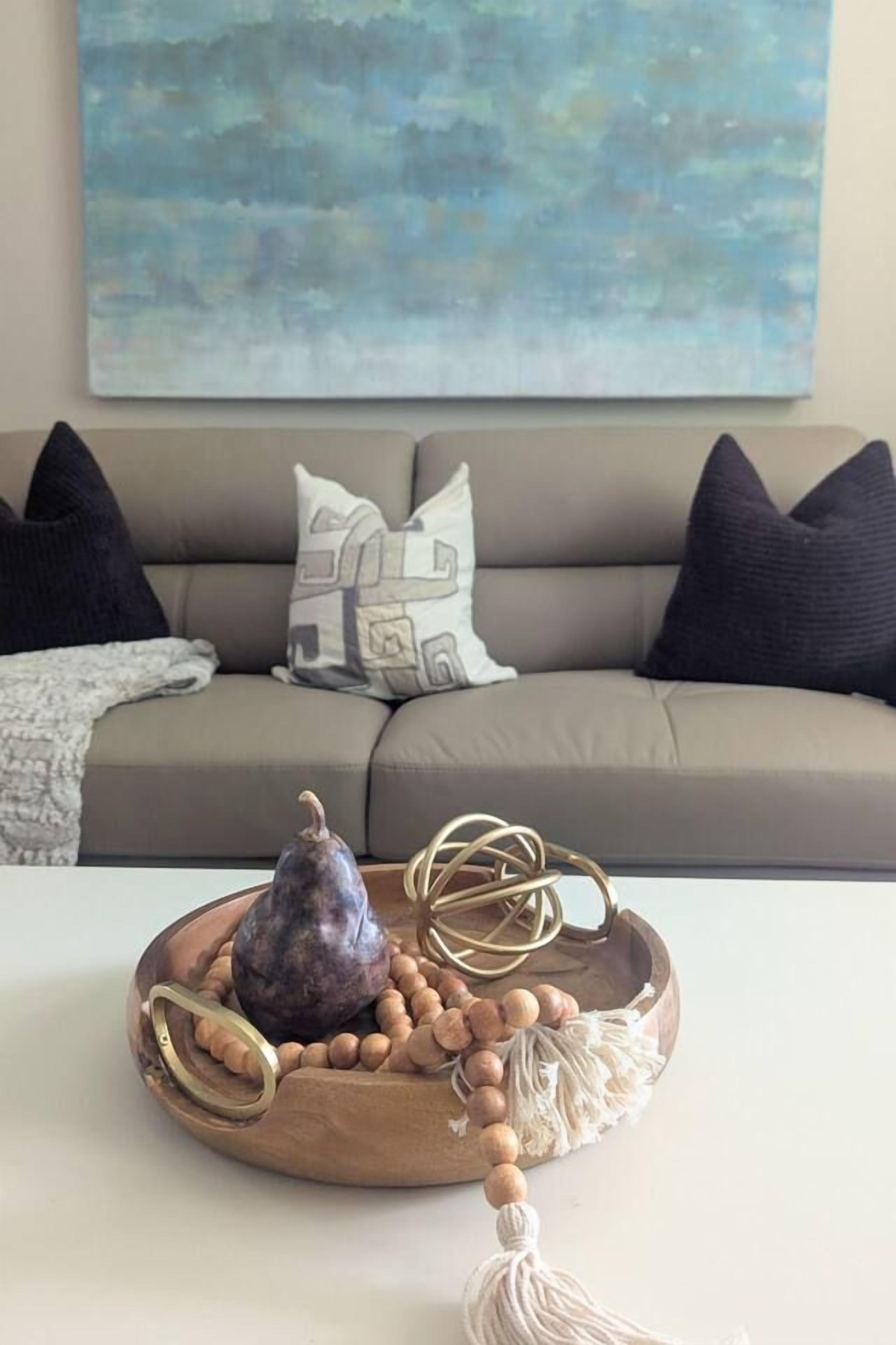 A living room with a sofa, a decorative tray with a fruit, a macrame piece, and a wall painting.