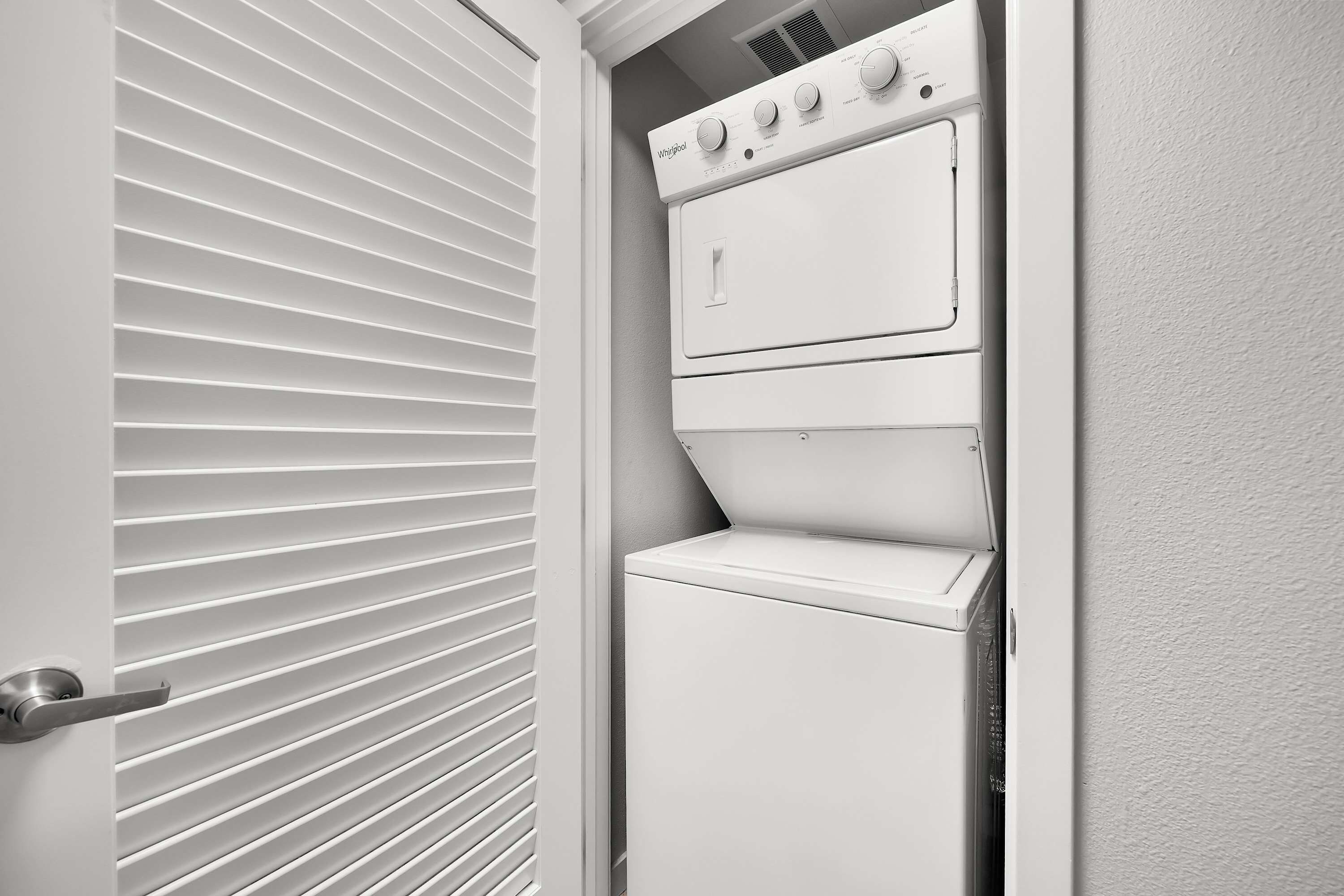A white laundry room with a built-in washer and dryer, a door with a handle, and a vent above the dryer.
