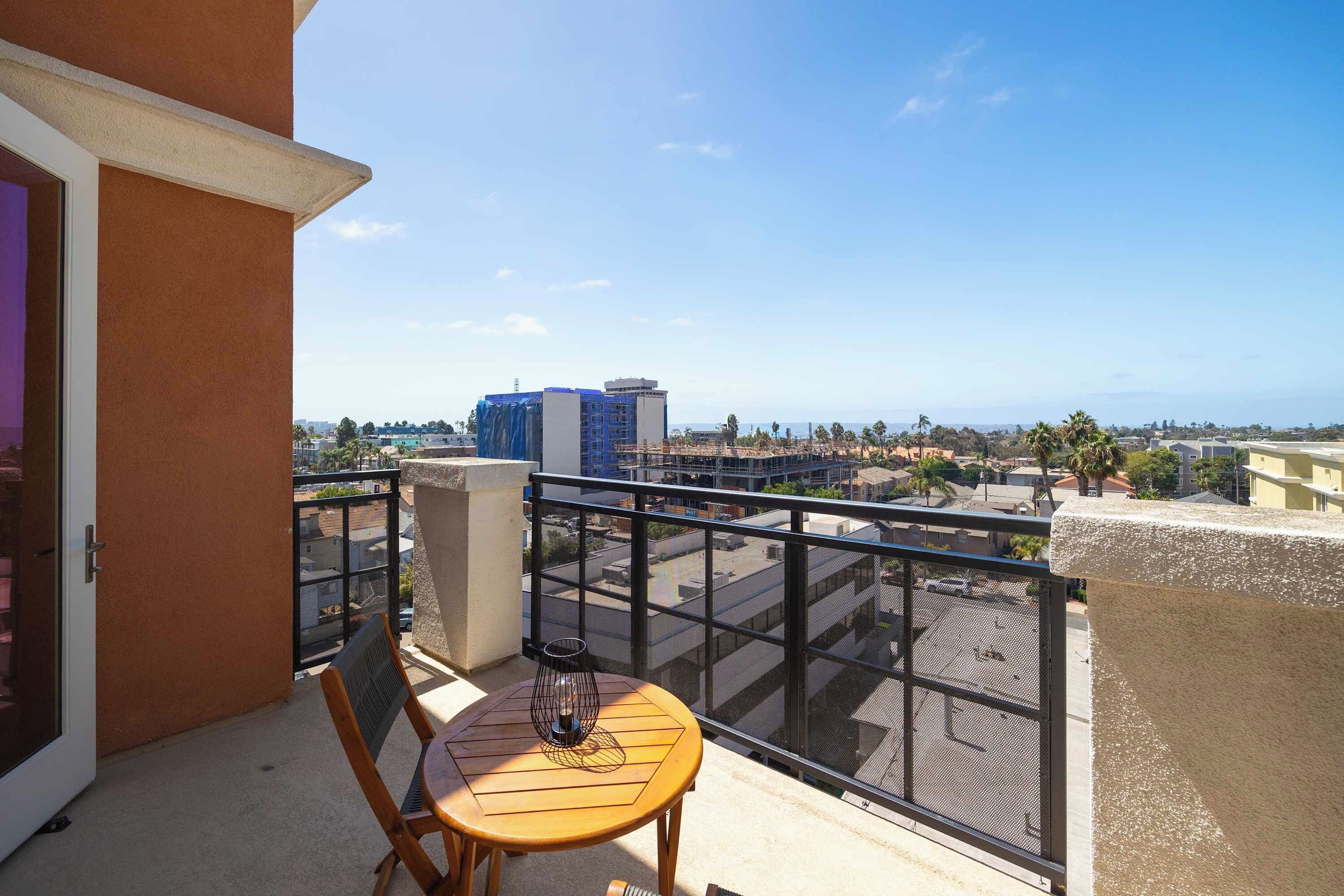 A balcony with a table and chair, overlooking a cityscape with buildings and palm trees.