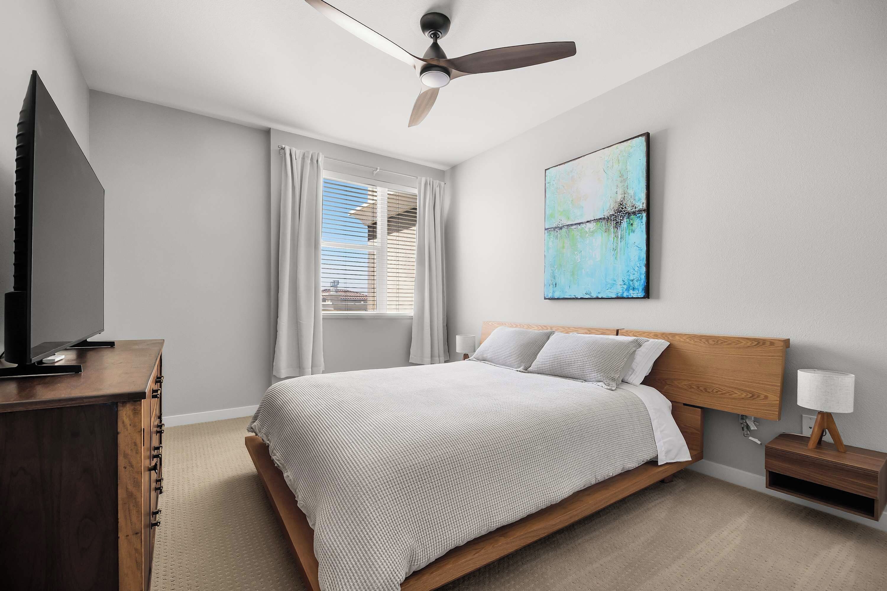 A hotel room with a bed, a TV, a ceiling fan, a painting, and a nightstand with a lamp.