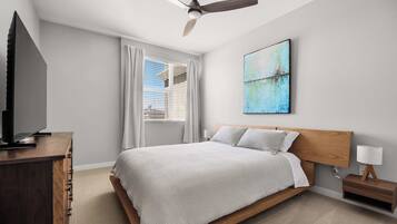 A hotel room with a bed, a TV, a ceiling fan, a painting, and a nightstand with a lamp.