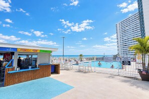 A beachside pool area with a concession stand, a fenced swimming pool, and a tall building in the background. - Golden Sands 101 (Ocean City)