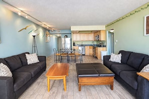 A living room with two black sofas, a wooden coffee table, and a dining table with chairs. There is a kitchen in the background with wooden cabinets and a microwave. - Golden Sands 101 (Ocean City)