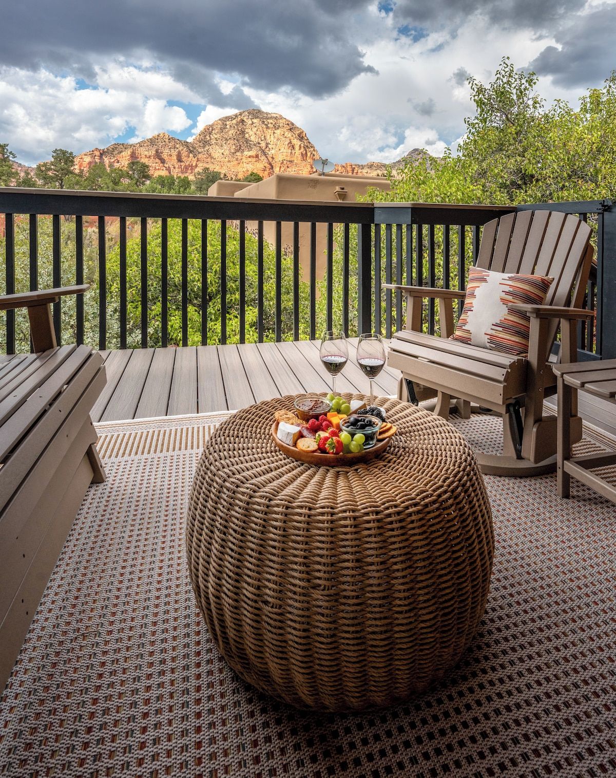 A wooden deck with a round woven ottoman, two chairs, and a table set with snacks and wine glasses.