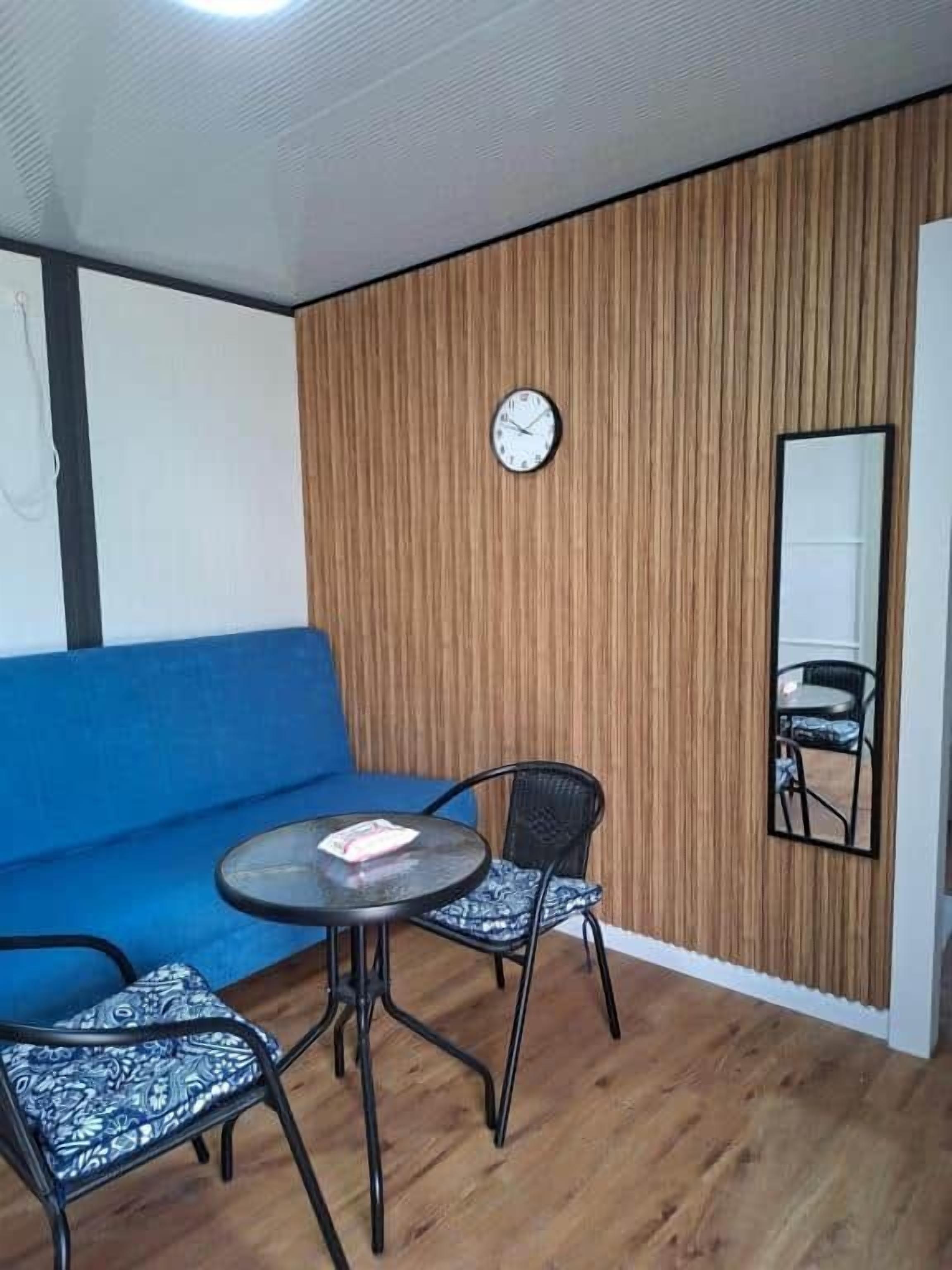 A room with a blue sofa, a round table with two chairs, a wooden wall panel, a clock, and a mirror.