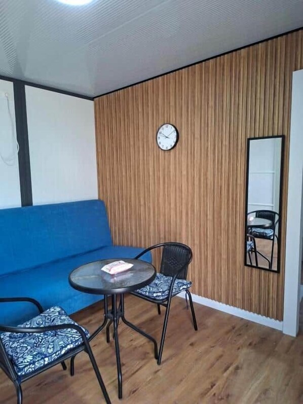 A room with a blue sofa, a round table with two chairs, a wooden wall panel, a clock, and a mirror.
