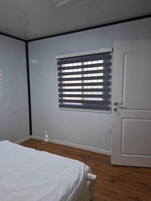 A bedroom with a bed, a window with blinds, a door, and a wooden floor.