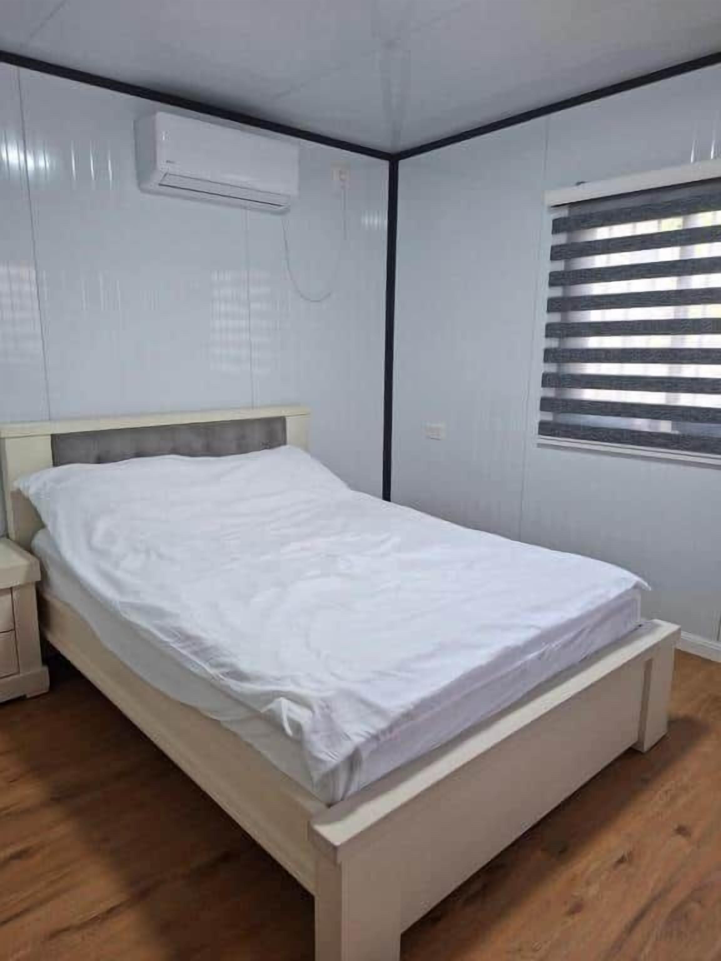 A single bed with a white mattress and headboard, a nightstand, a wall-mounted air conditioner, and a window with blinds.