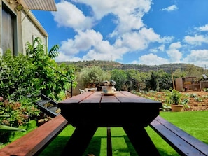 A wooden picnic table with a cup and saucer in the center, surrounded by a well-maintained garden with various plants and a clear sky. - Spacious Tiny House – Caravan Style (Majd al-Krum)