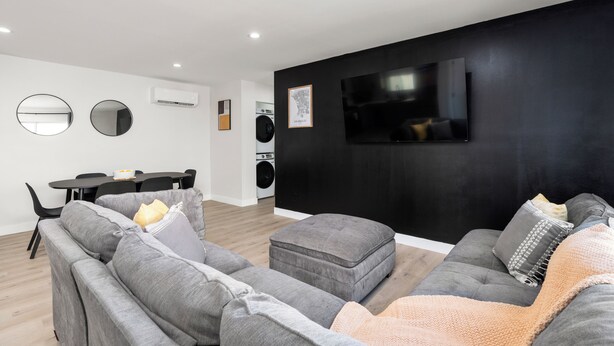 Een moderne woonkamer met een grijze zithoek, een flatscreen televisie aan een zwarte muur en een eethoek met een tafel en stoelen.